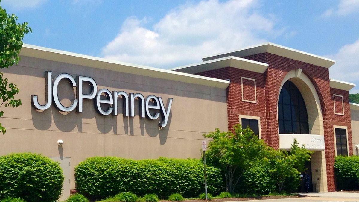 With puffy clouds in a blue sky, bushes surround a store with the name "JC Penney" in white letters. The store entrance is a brick arch.