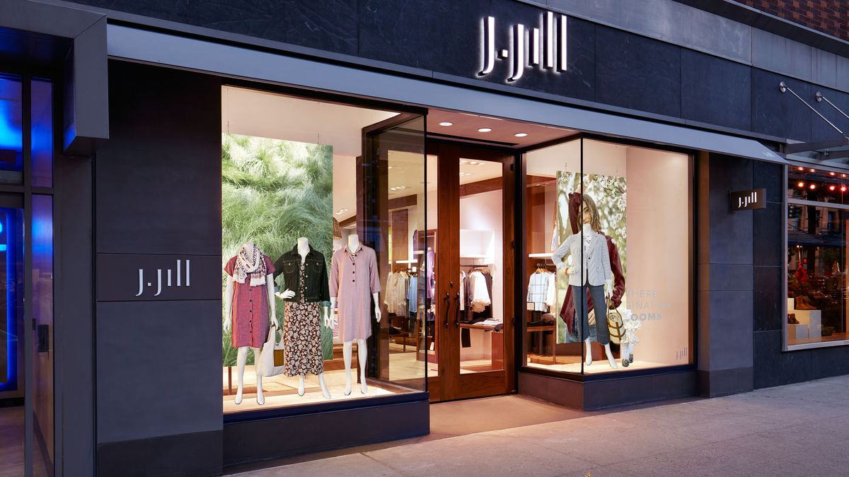 Lit-up store windows show mannequins wearing apparel, including dresses on the left and jeans and a jacket on the right. The banner name "J.Jill" is above and to each side of the door.