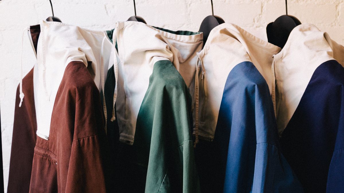 Four anoraks, (brown, green, blue and navy) hang on a black rack. their collars and the background are cream-colored.