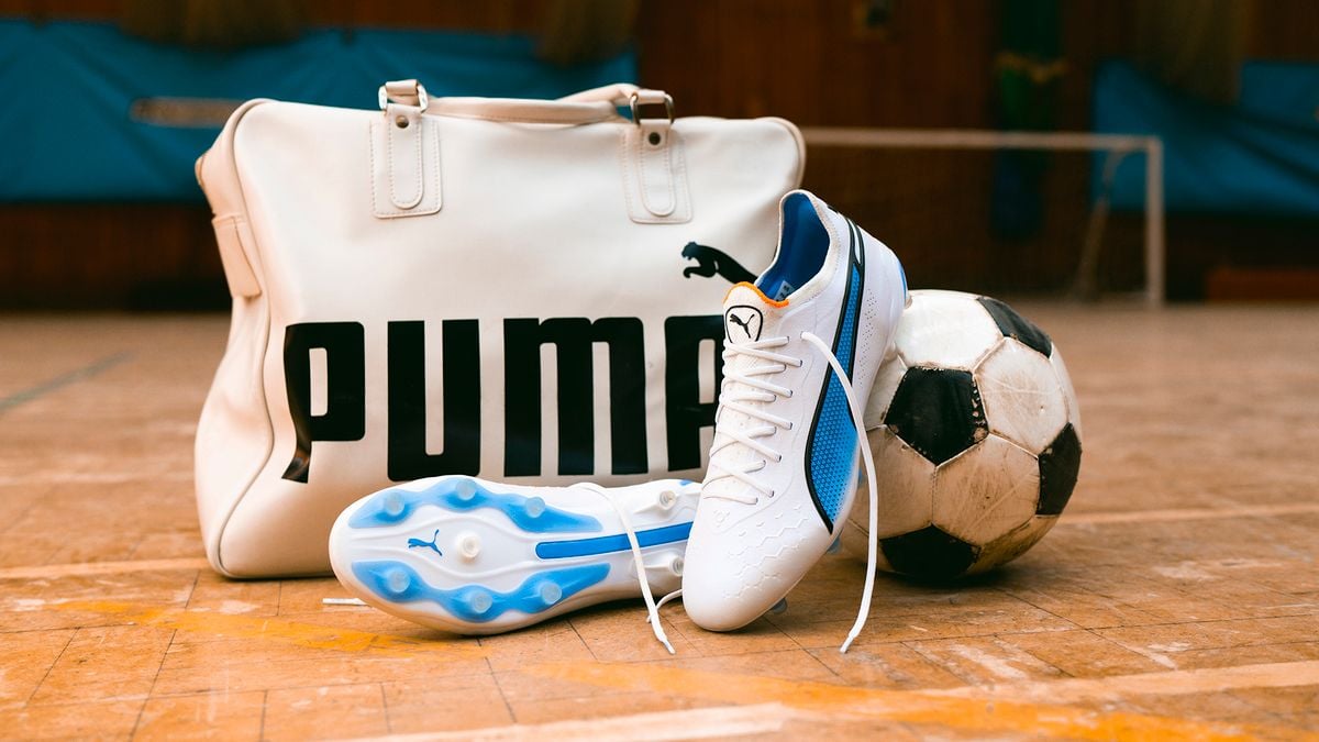On the brown tile floor of an indoor soccer pitch, a white scuffed up gear bag with the word PUMA sits behind a pair of blue and white soccer cleats. One cleat leans against a soccer ball.