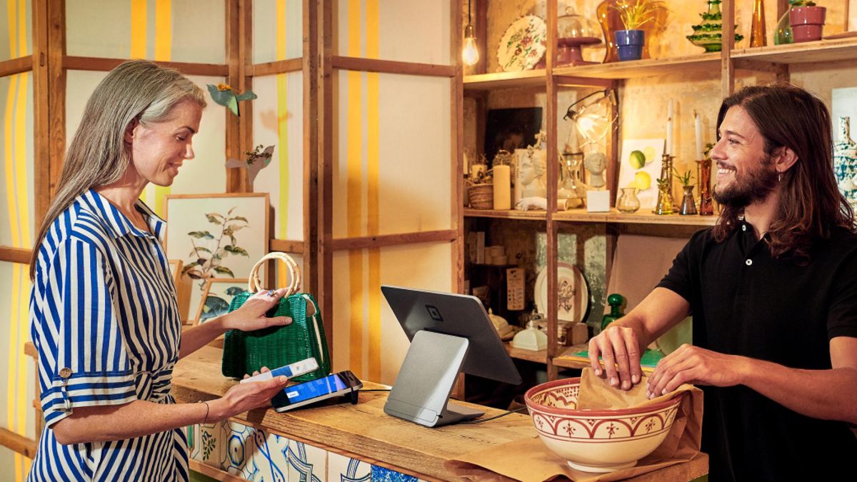 A customer uses a mobile device to make a transaction while a cashier looks on with a Square device in between them.