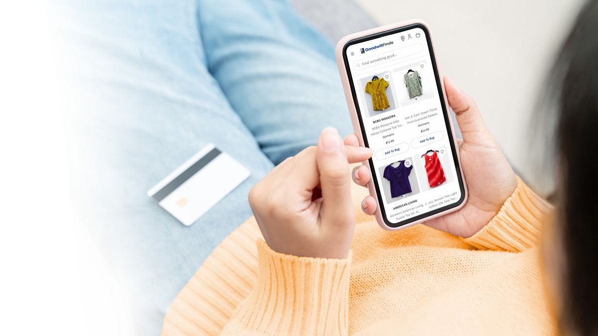 A person wearing light blue jeans and a yellow sweater has a credit card handy as they browse a selection of clothing on their phone.
