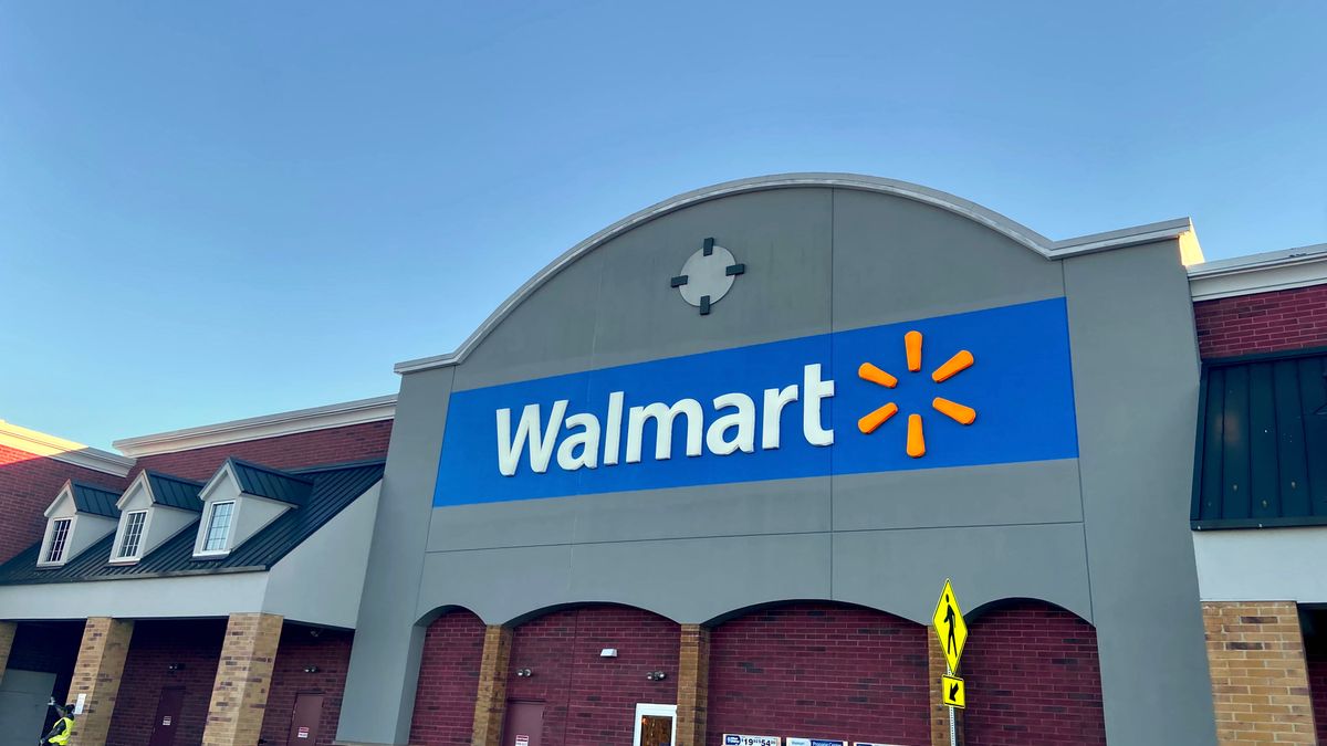 The curved banner of a Walmart store, the brand name in white letters and the characteristic yellow sunburst logo to the right, against a blue sky.