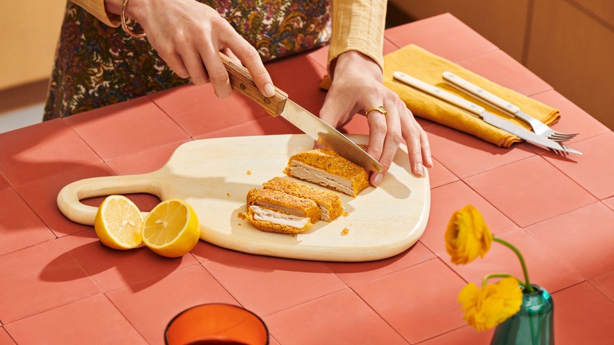 A person slices a Meati Crispy Cutlet on a white cutting board that sits on a coral-colored counter.